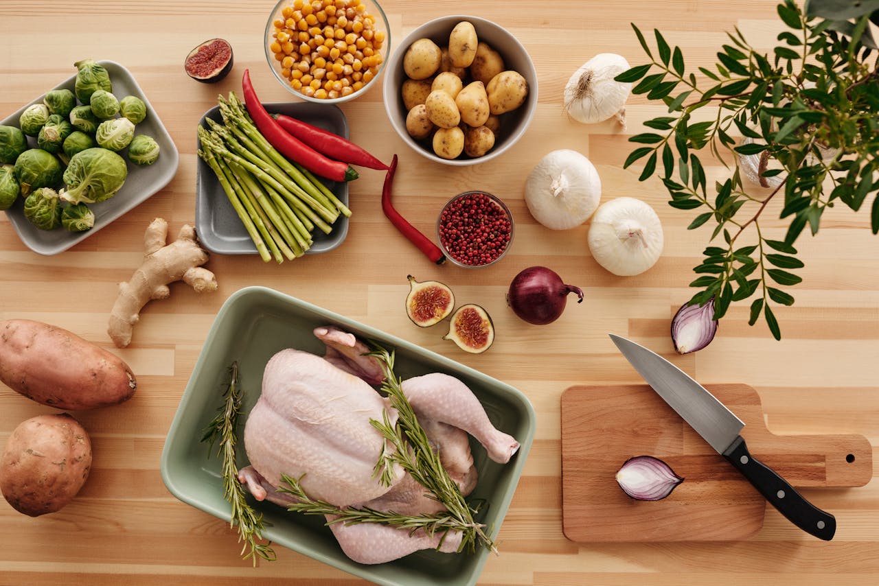 Top view of fresh ingredients for cooking on a wooden surface, featuring chicken, vegetables, and herbs.